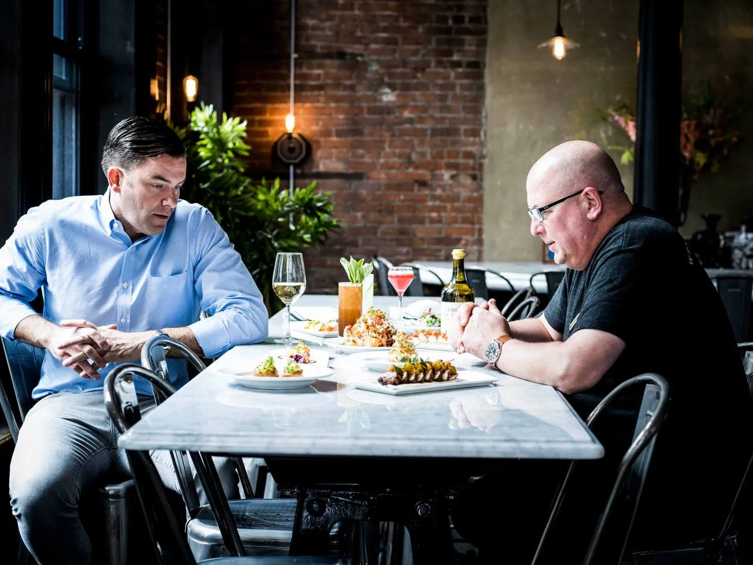 Founder Dave Kwiatkowski and Chef and Partner Marc Djozlija seated across a marble table spread with small plates and cocktails in the Wright & Company dining room