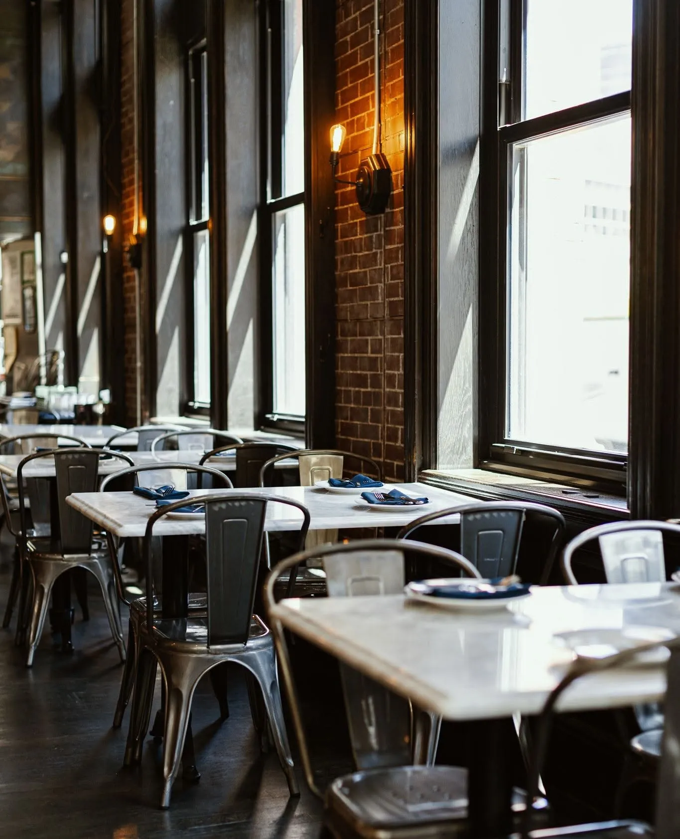 Marble tables and metal chairs by the tall windows at Wright and Company, set with navy napkins over exposed brick