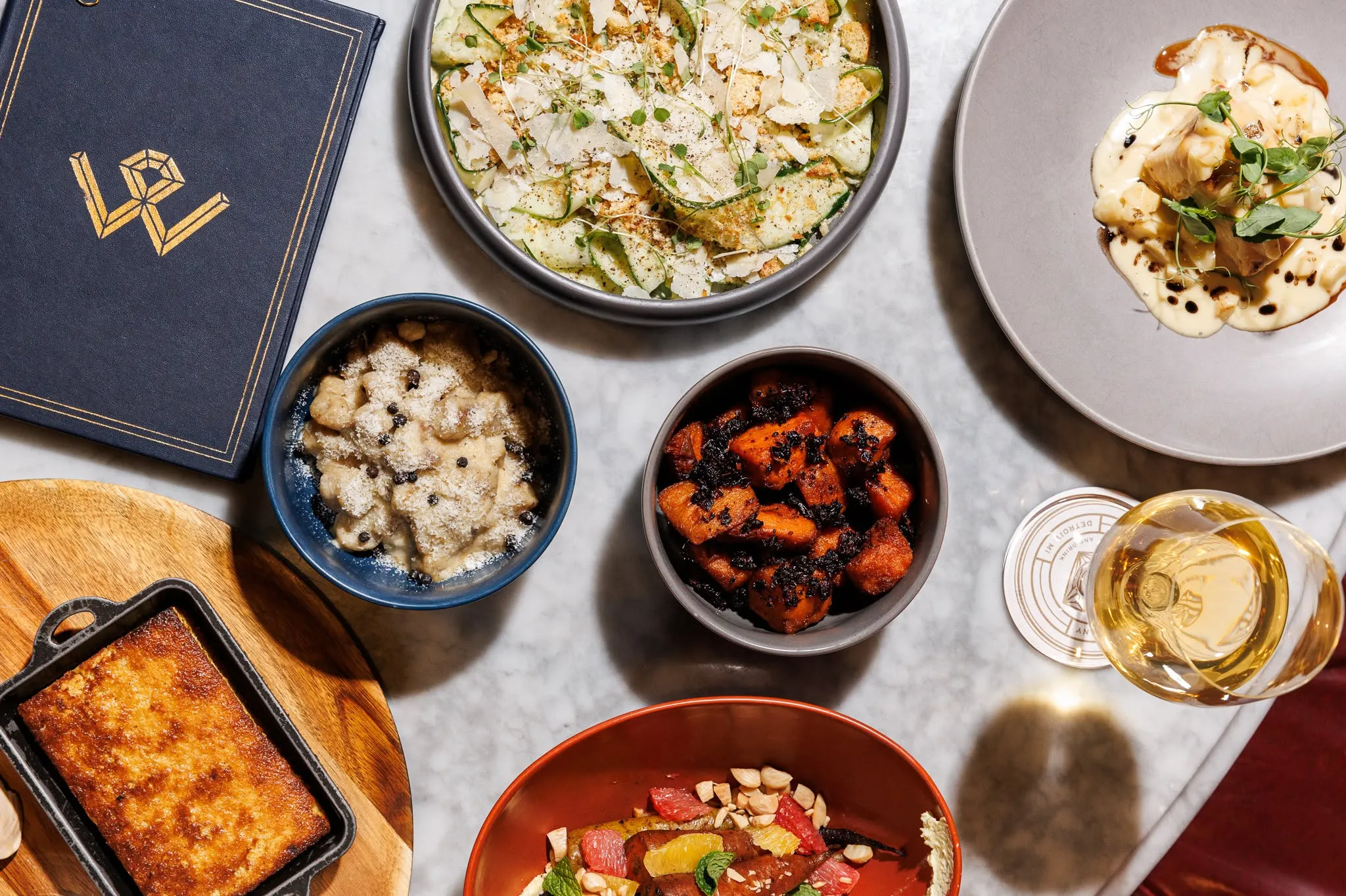 An overhead spread of shared plates on a marble table at Wright and Company: a shaved zucchini and parmesan salad, gnocchi, roasted sweet potatoes, cornbread, and a glass of white wine beside a gold-monogram menu