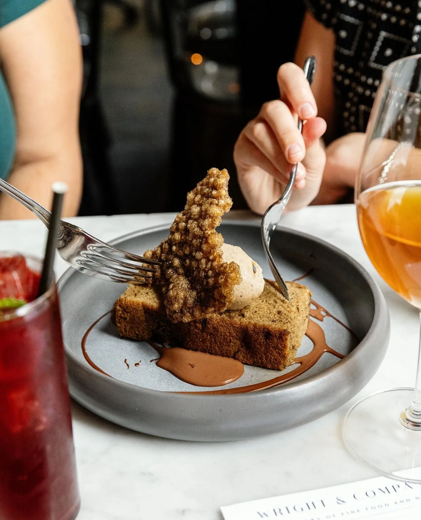 Two guests sharing dessert at Wright and Company: a slice of spice cake with a mousse quenelle and caramelized brittle over chocolate sauce, forks in hand, beside a cocktail and a glass of wine and a Wright and Company menu card.